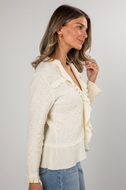 Woman wearing a textured white cardigan over a blouse with ruffled details on a gray background