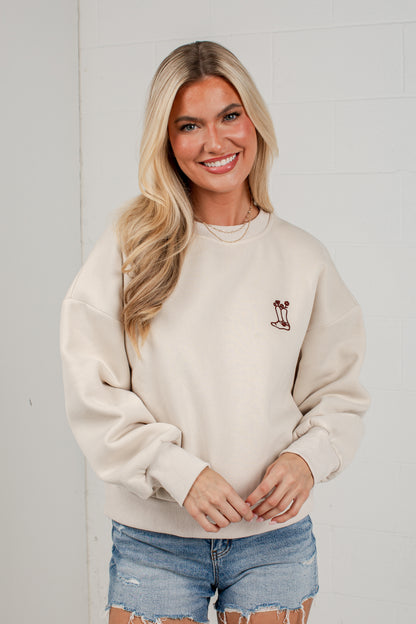 A smiling blonde woman wearing a cream-colored crewneck sweatshirt with a small embroidered palm tree graphic on the left chest, paired with distressed denim shorts, standing against a white brick wall.