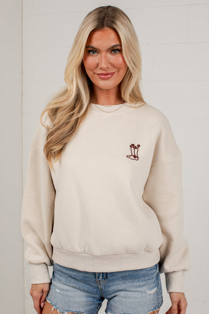 A blonde woman wearing a cream-colored crewneck sweatshirt with a small embroidered palm tree graphic on the left chest, paired with distressed denim shorts, standing against a white brick wall.