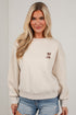 A blonde woman wearing a cream-colored crewneck sweatshirt with a small embroidered palm tree graphic on the left chest, paired with distressed denim shorts, standing against a white brick wall.