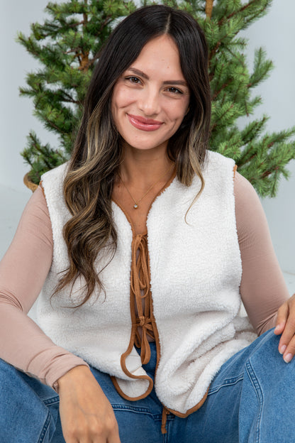 Woman wearing a white fleece vest with a brown leather strap in front of a Christmas tree.