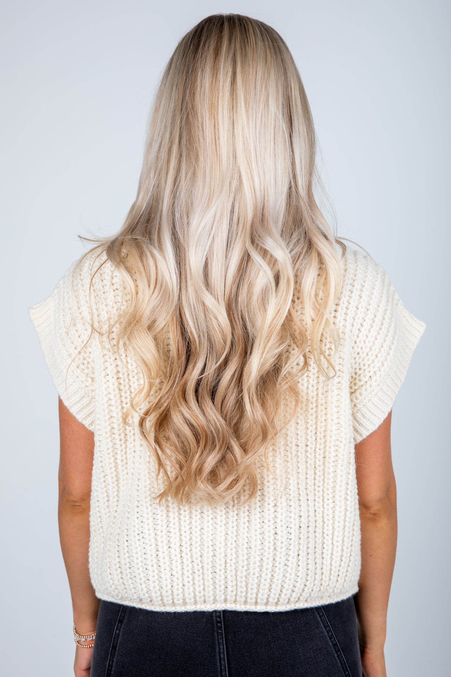 Person wearing a white knit sleeveless top with long, wavy blonde hair on a plain background