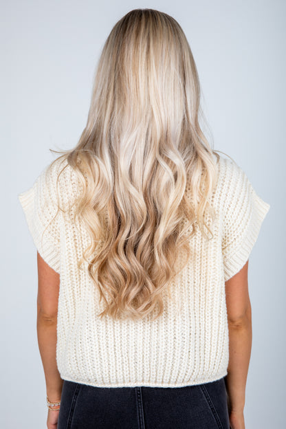 Person wearing a white knit sleeveless top with long, wavy blonde hair on a plain background