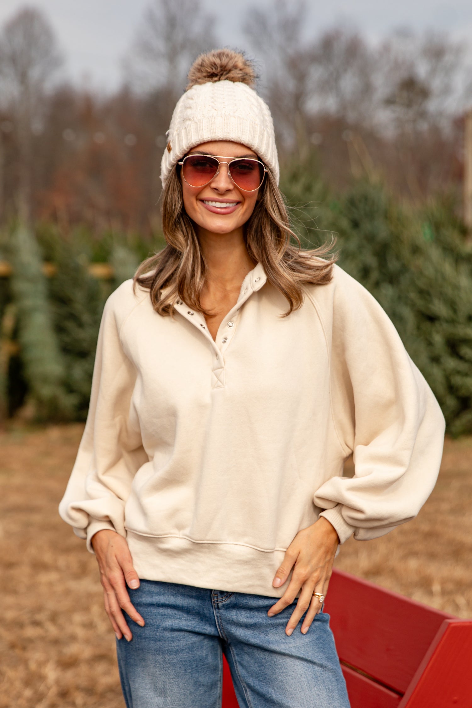 Woman wearing a cream sweater, sunglasses, and a white beanie in a Christmas tree lot.
