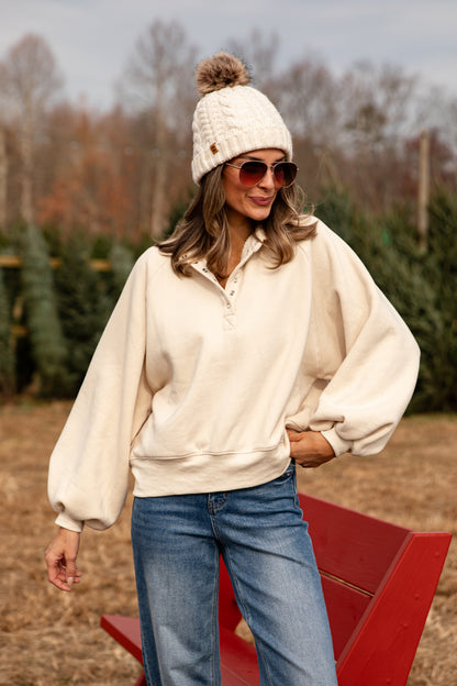 Woman wearing a cream sweater, blue jeans, and a white knit beanie with a pom-pom, standing in a field with trees.