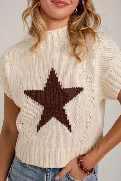 A woman wearing a cream-colored knit sweater with a large brown star motif on the chest, paired with blue jeans.