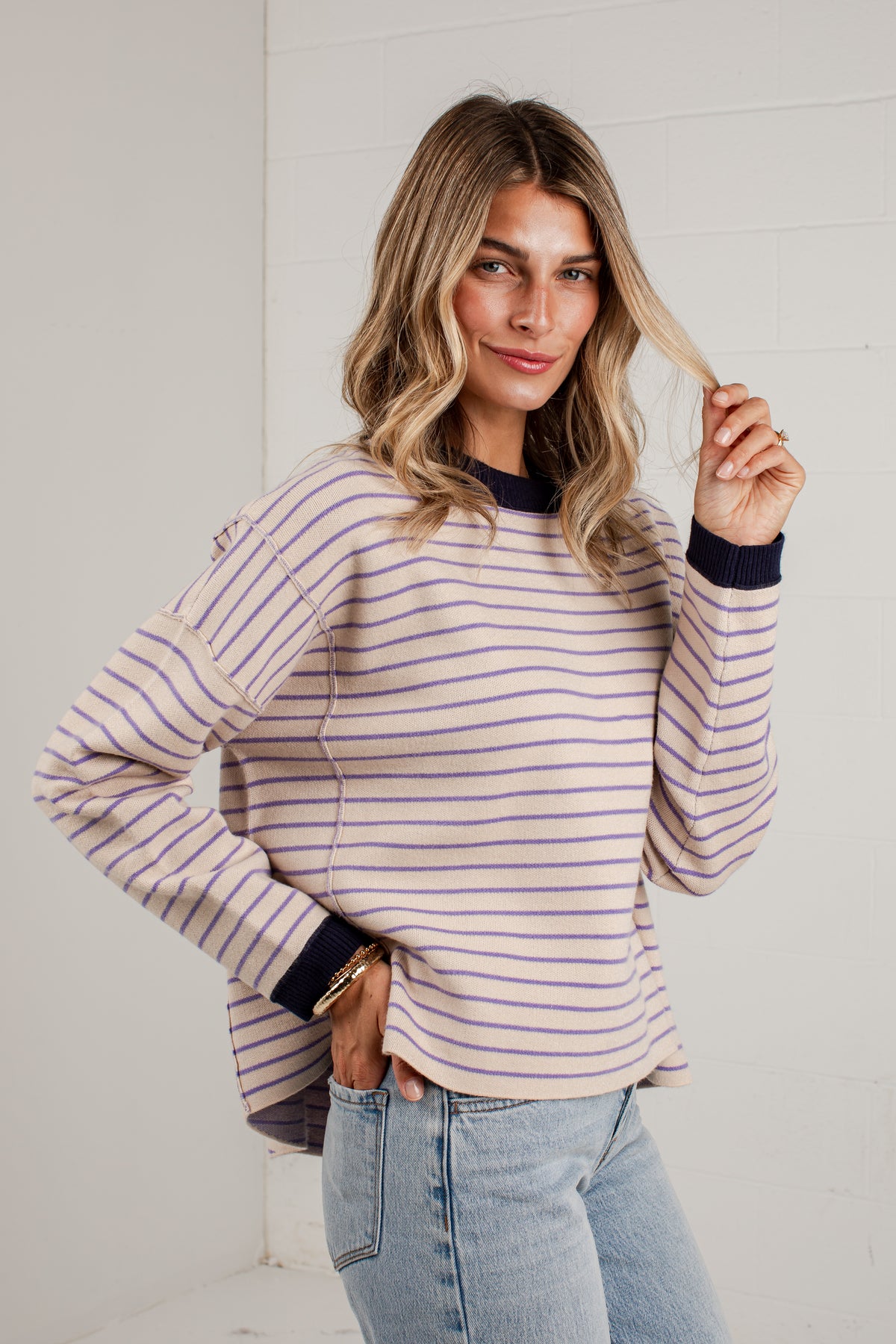 A woman wearing a long-sleeve knit sweater with thin lavender and beige horizontal stripes, featuring dark navy ribbed trim at the crew neck, cuffs, and hem, paired with light-wash blue jeans.