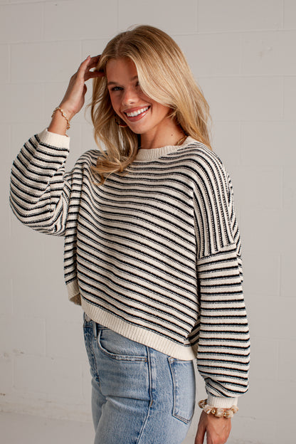 A woman wearing a black and cream striped knit sweater with long sleeves and a cropped hem, paired with light blue jeans, standing against a white brick wall.