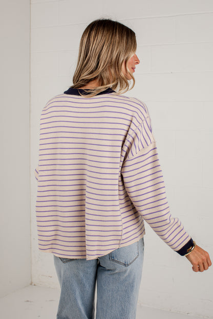 A woman shown from the back wearing a light pink and navy blue horizontally striped long-sleeve top with light-wash blue jeans, standing against a white brick wall.