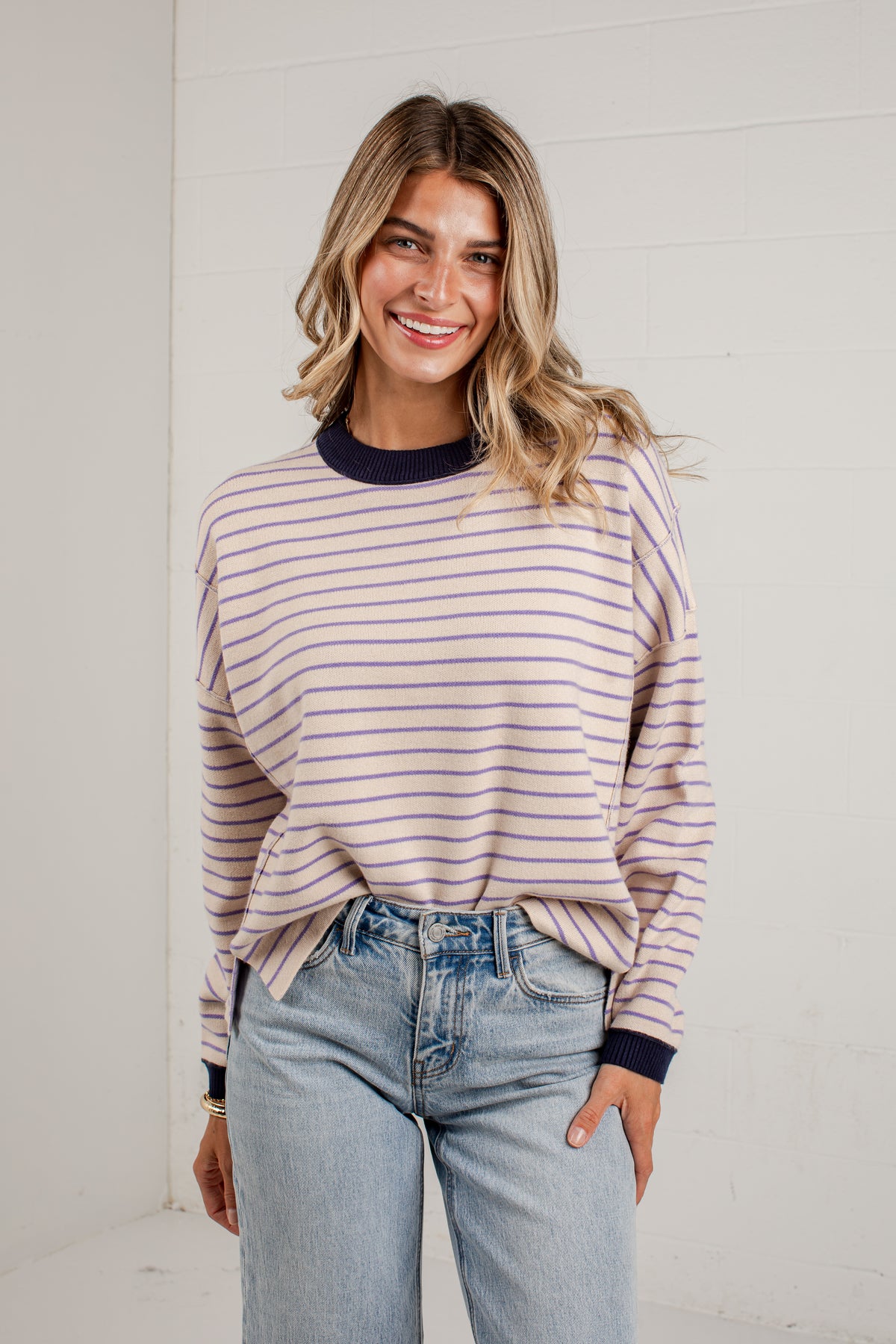 A woman wearing a light pink and navy blue horizontally striped long-sleeve knit top with a dark navy crew neck, paired with light-wash blue jeans, standing against a white paneled wall.