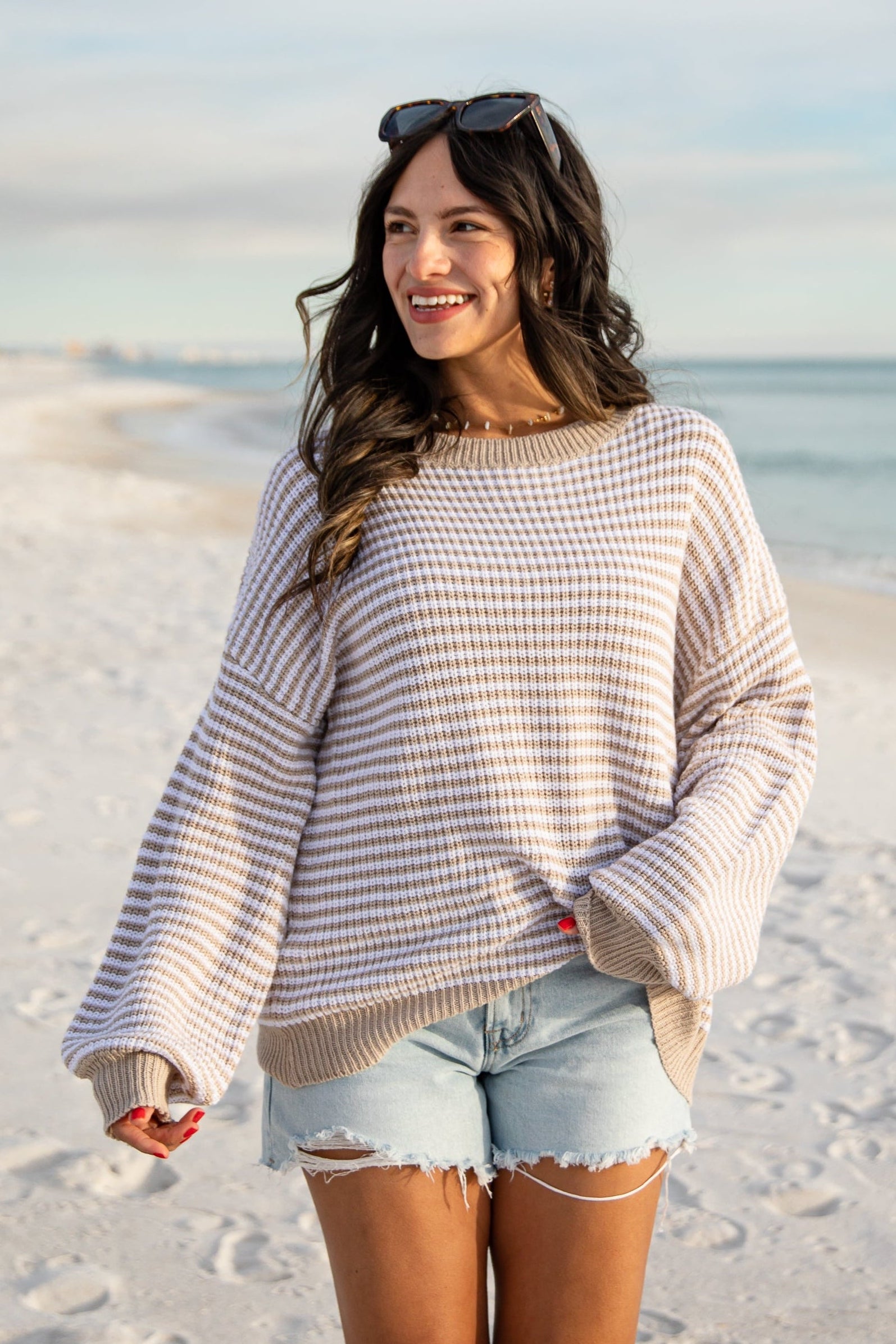 Woman standing on a sandy beach wearing a striped sweater and denim shorts.