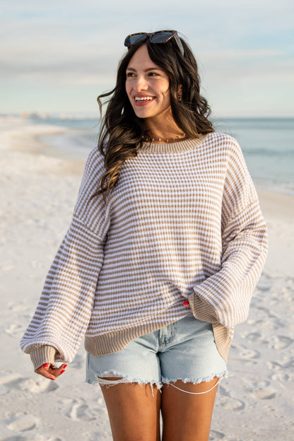 Woman standing on a sandy beach wearing a striped sweater and denim shorts.