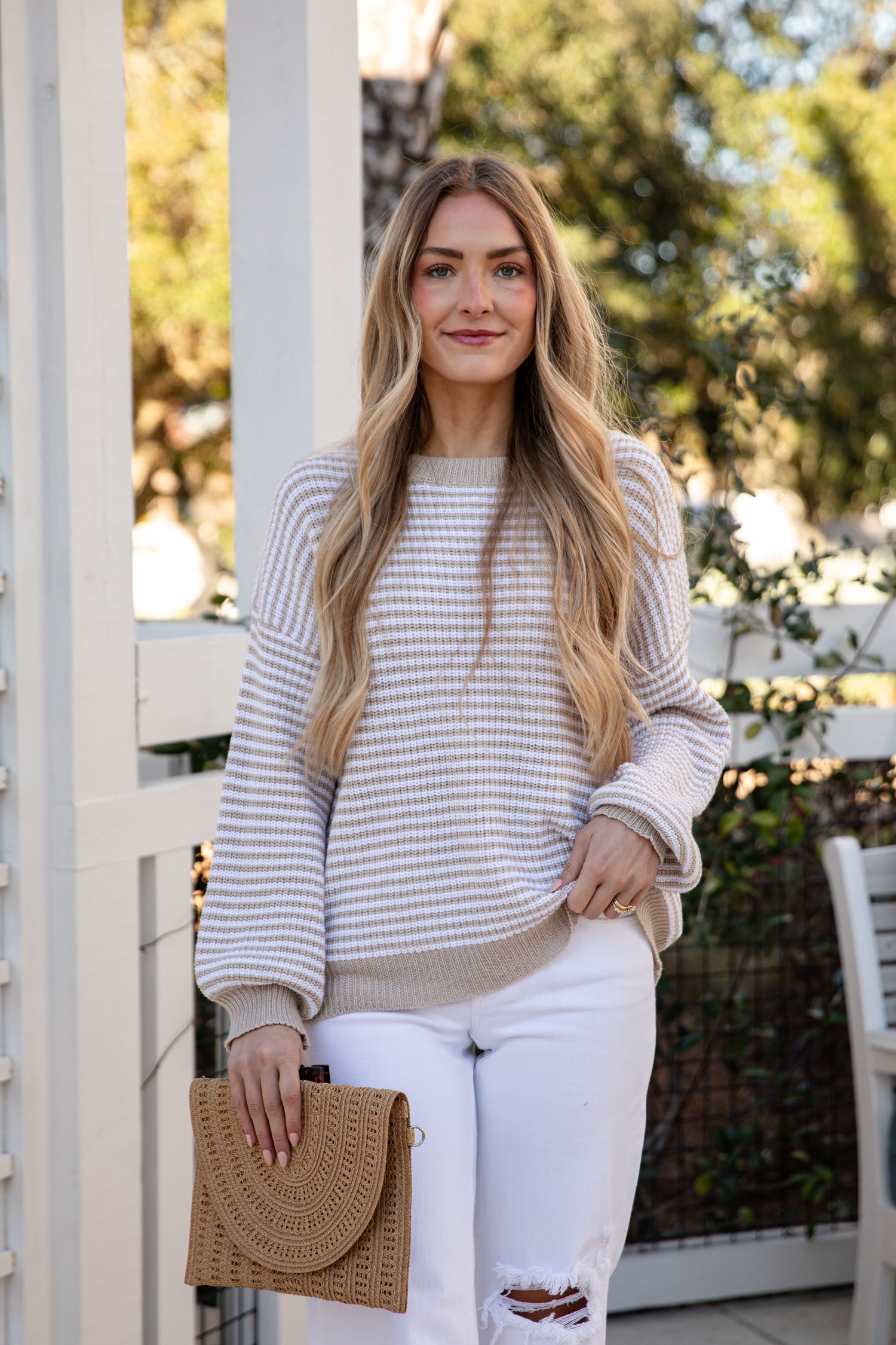Woman standing outdoors holding a beige handbag, wearing a striped sweater and white pants.