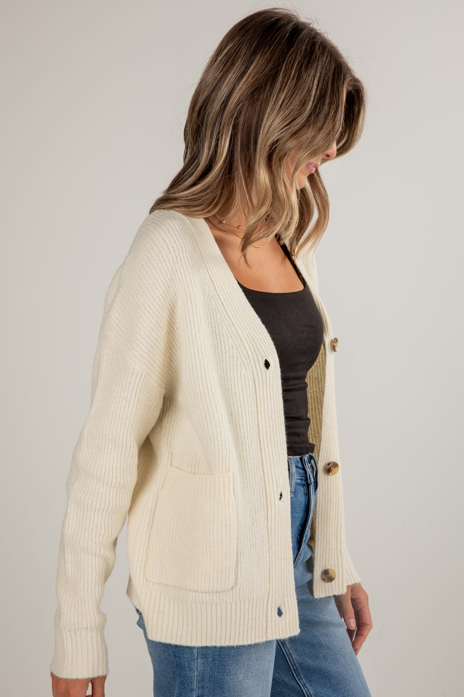 Woman wearing a cream cardigan over a black top and blue jeans on a plain background