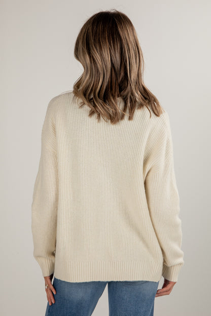 Person wearing a beige sweater and blue jeans against a plain background