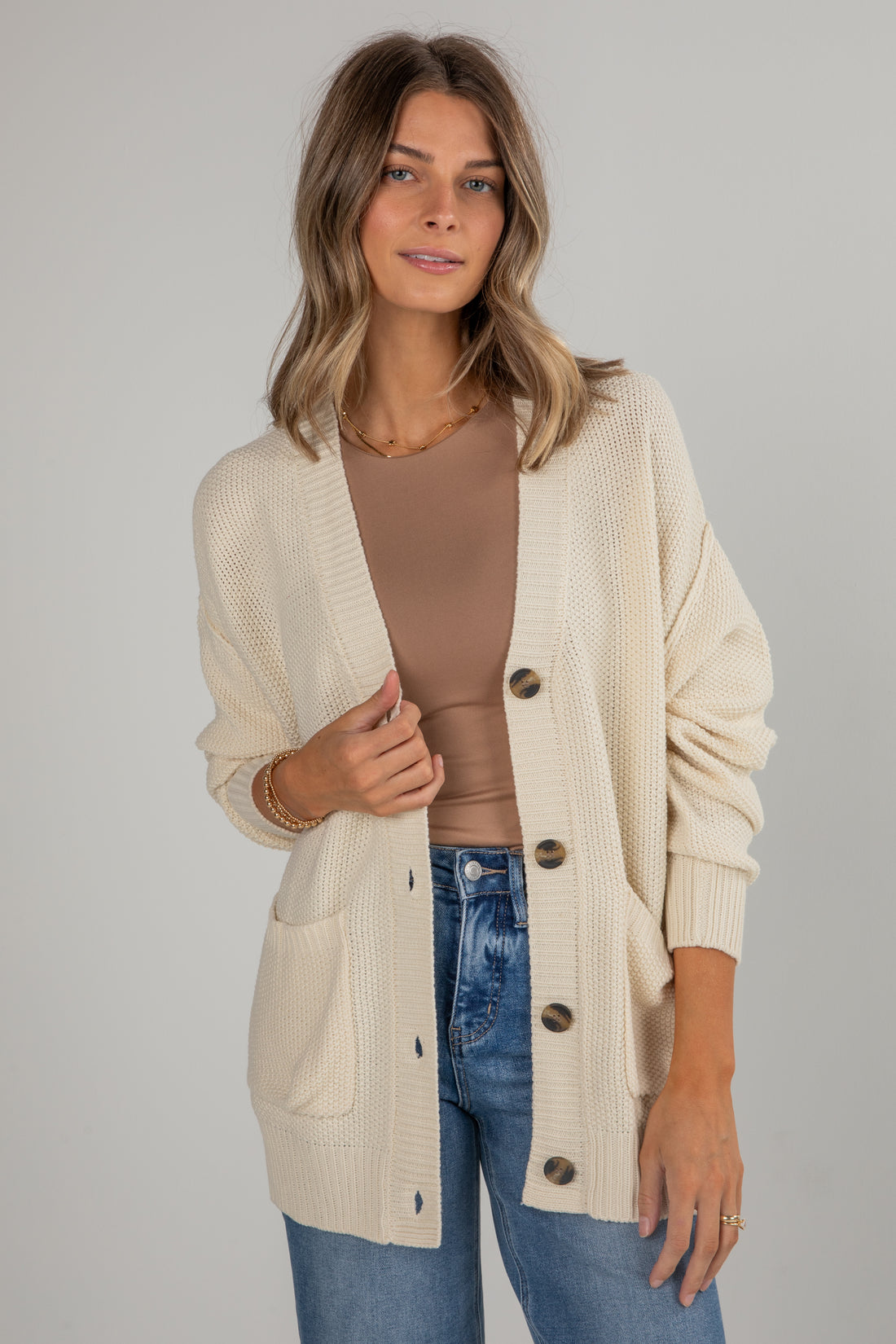 Woman wearing a beige cardigan over a brown top and blue jeans on a gray background