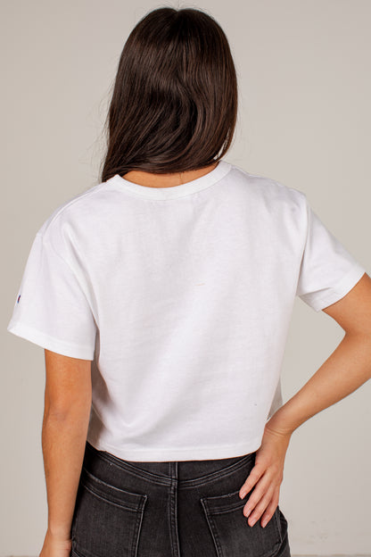 A woman shown from behind wearing a white short-sleeve cropped T-shirt and dark gray jeans, standing against a plain light beige studio background.