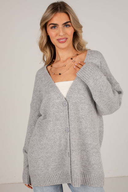 A woman wearing a light gray oversized knit cardigan with a V-neck and front buttons, layered over a white top and light blue jeans, against a plain light gray studio background.