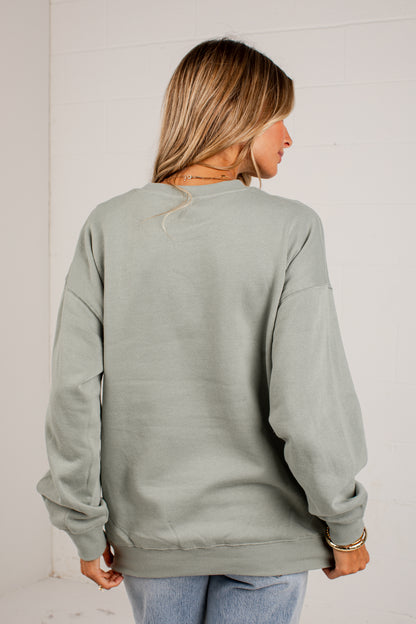 A woman viewed from the back wearing an oversized sage green crewneck sweatshirt and light blue jeans, standing against a white paneled wall.