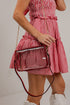 A person wearing a red and white gingham dress carries a clear rectangular crossbody bag with red trim, a gold chain strap, and a red-and-black shoulder strap, against a plain light wall.