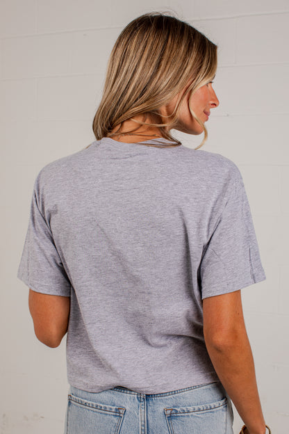 A woman shown from the back wearing a light heather gray short-sleeve t-shirt and light blue jeans, standing against a white brick wall.