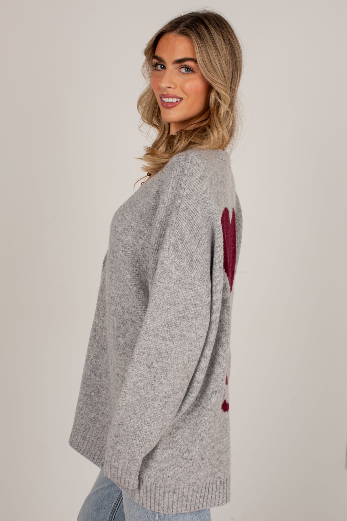 A woman wearing a light gray knit sweater with a burgundy heart design on the back, photographed against a plain light gray studio background.