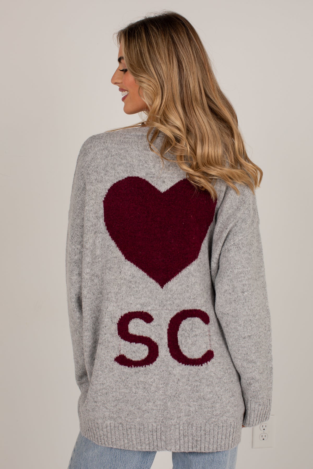 A woman shown from the back wearing a light gray knit sweater with a large dark red heart and the letters &