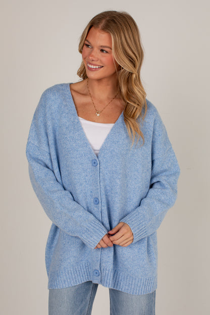 A woman wearing a light blue heathered knit cardigan over a white top and light blue jeans, standing against a plain beige studio background.