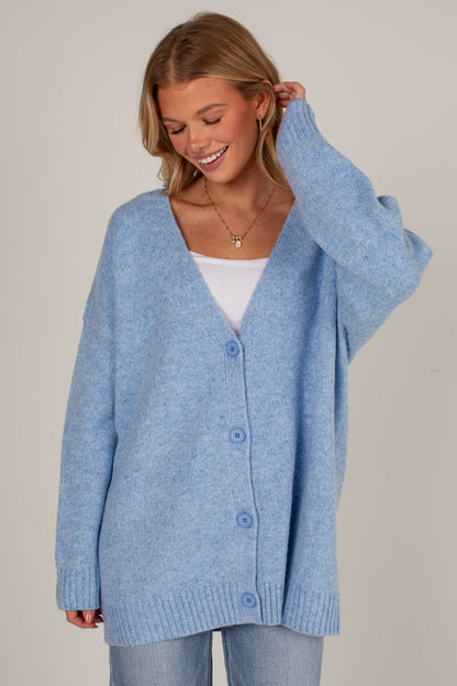 A woman wearing an oversized light blue knit cardigan with a white V-neck undershirt and light blue jeans, photographed against a plain light beige studio background.