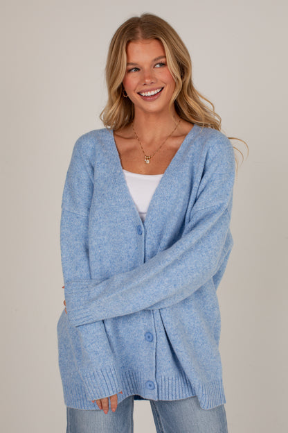 A smiling blonde woman wearing a light blue heathered knit cardigan over a white top and light blue jeans, photographed against a plain warm beige studio background.