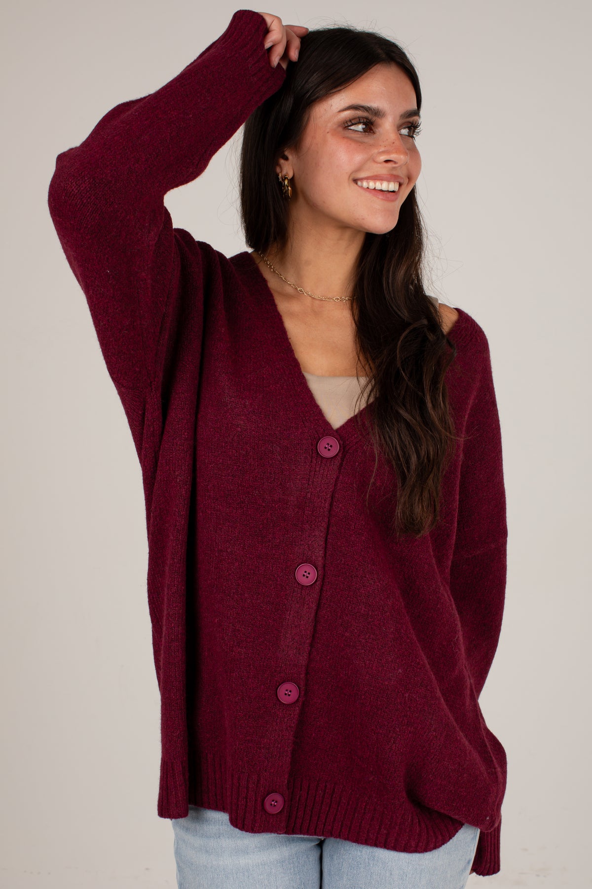 A woman wearing a maroon button-up knit cardigan sweater over a light beige top and light blue jeans, posing against a neutral light gray background.