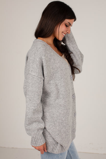 A woman wearing a light gray heathered knit cardigan sweater with a V-neck and button-front closure, paired with light blue jeans, photographed against a plain off-white wall.