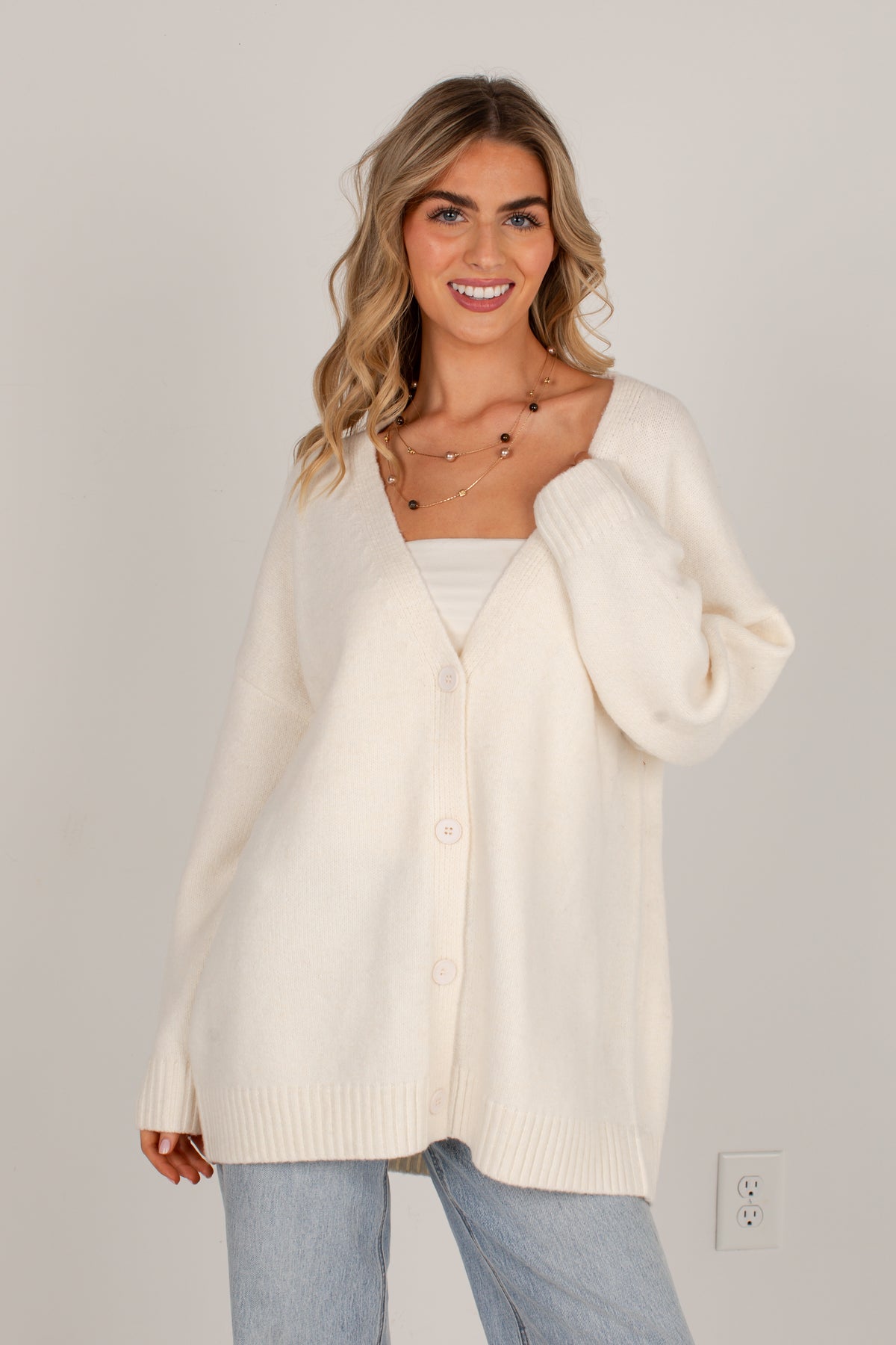 A woman wearing an oversized cream-colored knit cardigan sweater over a white top and light blue jeans, standing against a plain light gray wall.