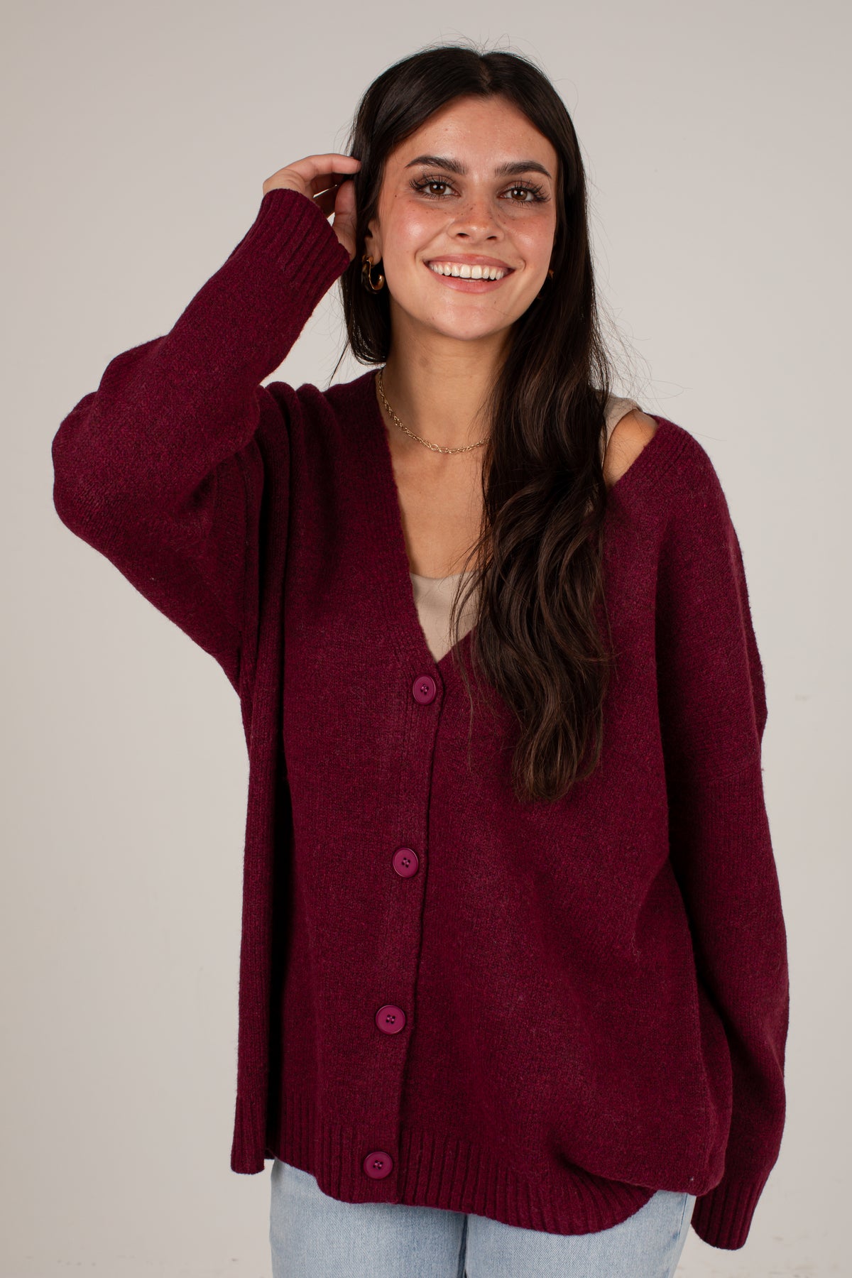 A woman wearing a burgundy knit cardigan with cold-shoulder detailing and pink buttons, paired with light blue jeans, against a plain light gray background.