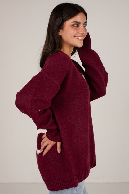 A woman wearing an oversized maroon knit sweater with a white graphic detail on the lower front, posing against a plain off-white background.
