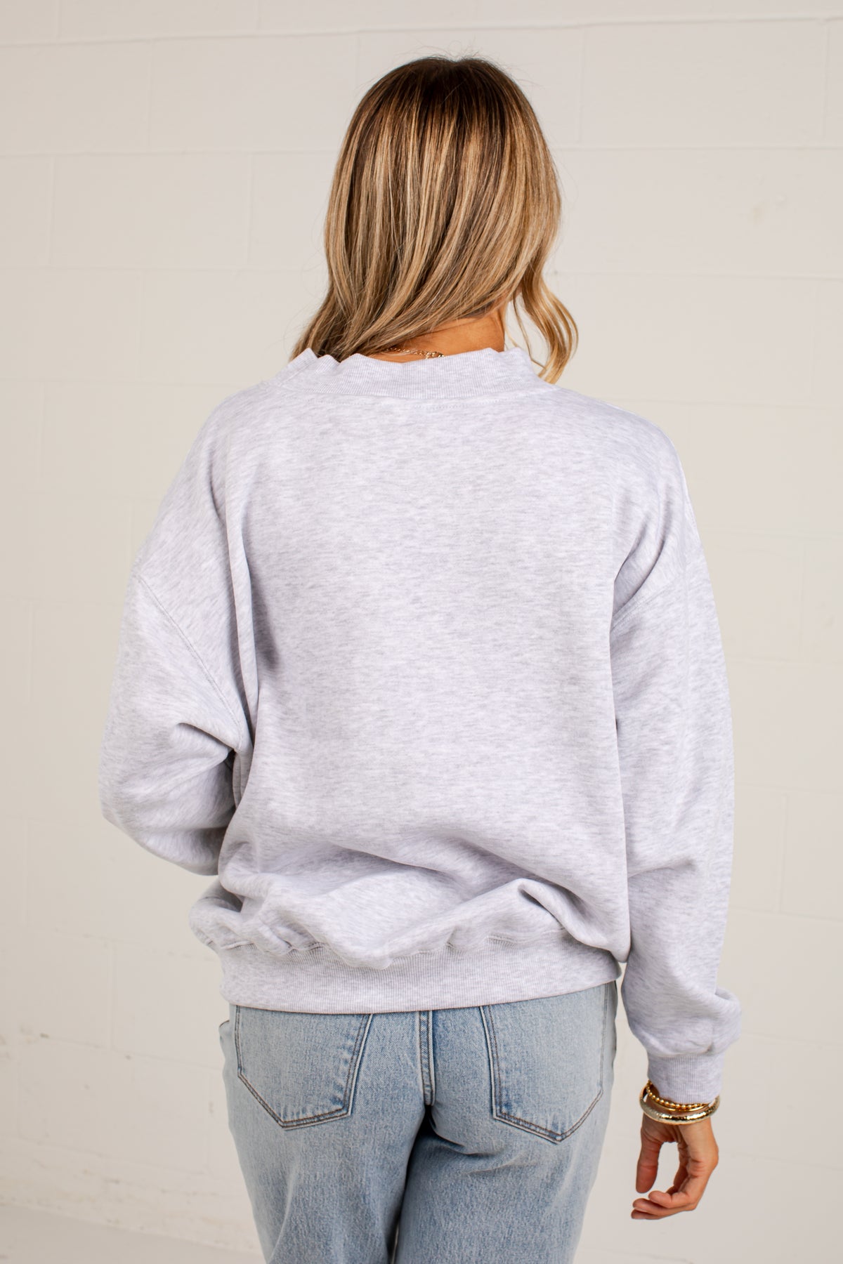 A woman viewed from behind wearing a light heather gray crewneck sweatshirt and light blue jeans, standing against a light beige wall.