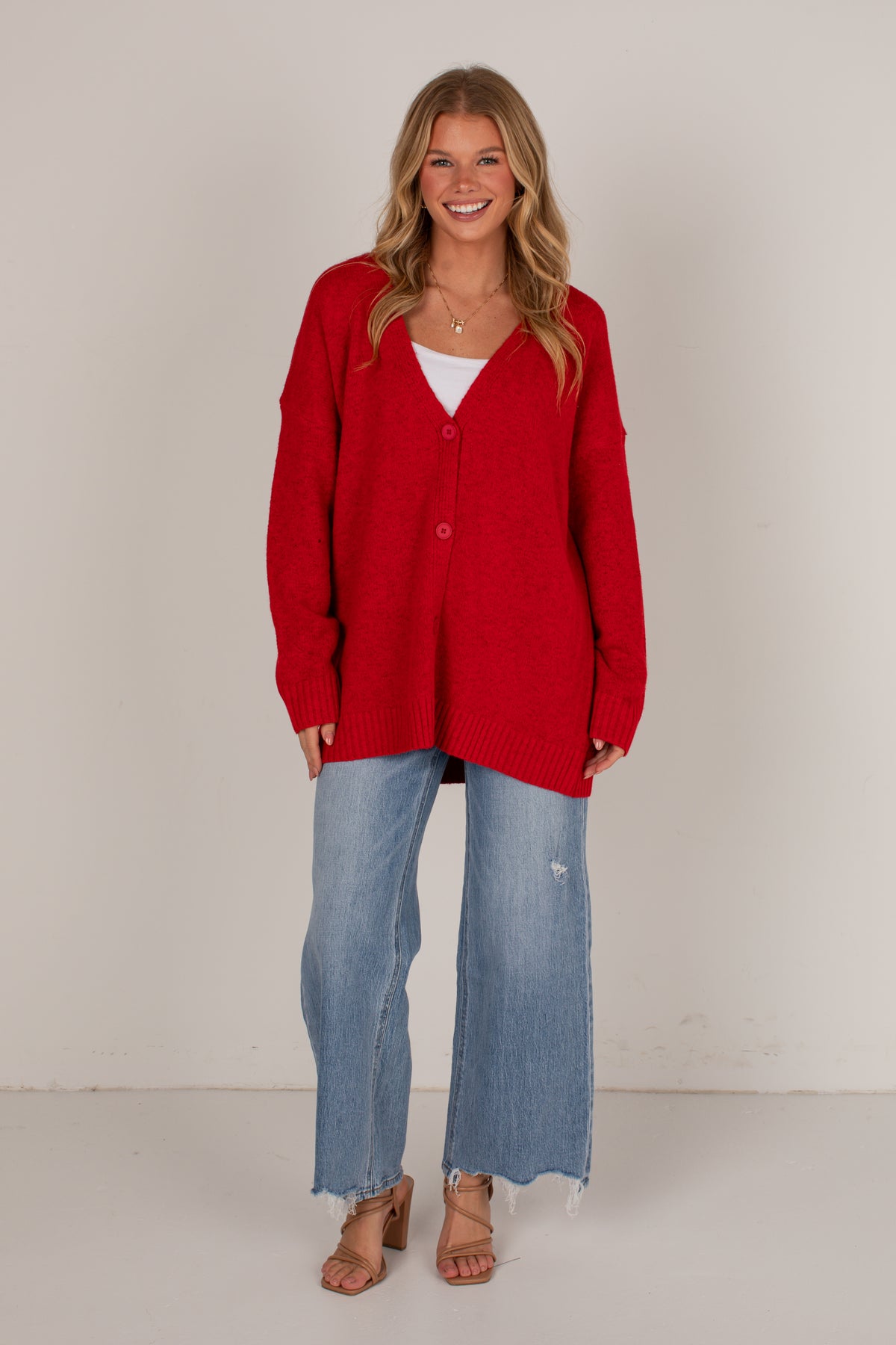 A woman modeling an oversized bright red knit cardigan over a white top, paired with light blue wide-leg jeans and brown heeled sandals, standing against a plain off-white studio background.