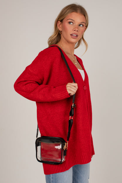 A woman wearing a bright red knit cardigan and light blue jeans, carrying a small black crossbody bag with a clear front pocket. She is photographed against a solid light gray studio background.