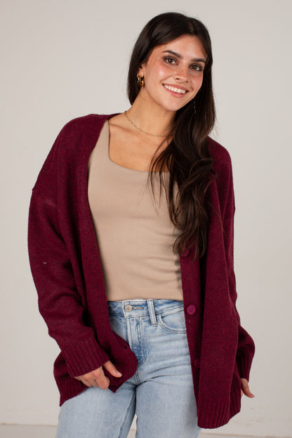 A woman wearing a maroon knit cardigan over a beige tank top and light blue jeans, standing against a plain light gray background.