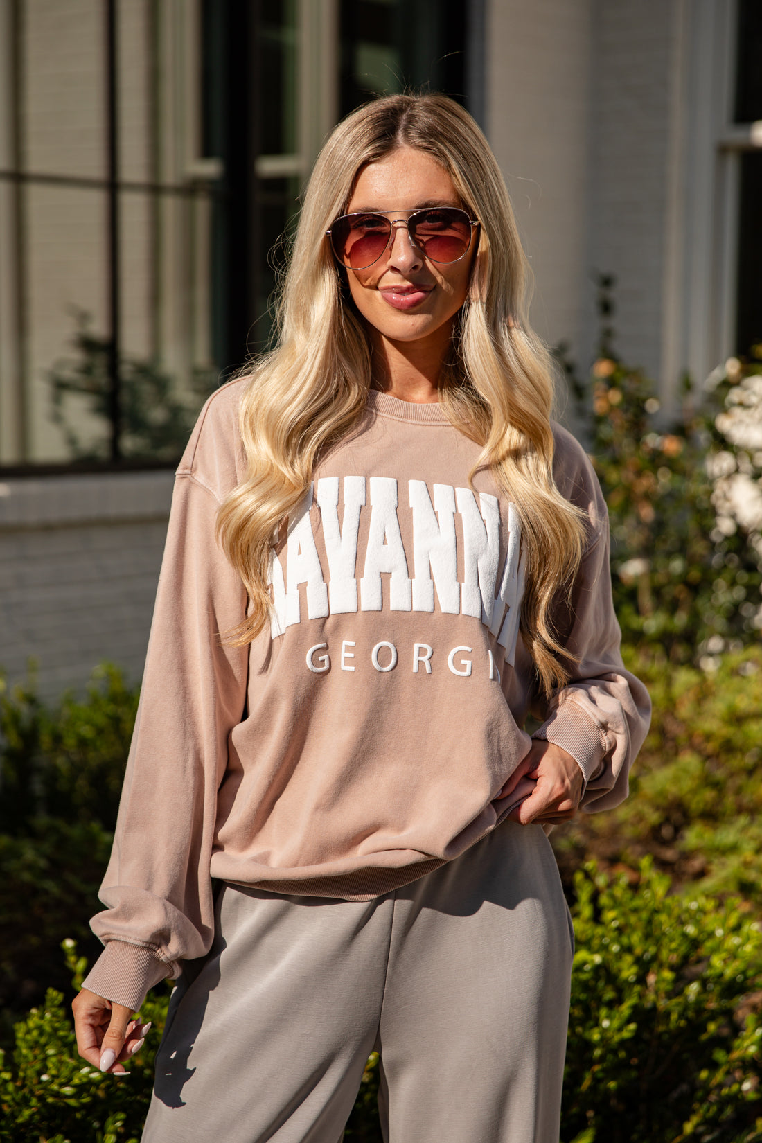Woman wearing a beige sweatshirt with text, sunglasses, and gray pants outdoors.
