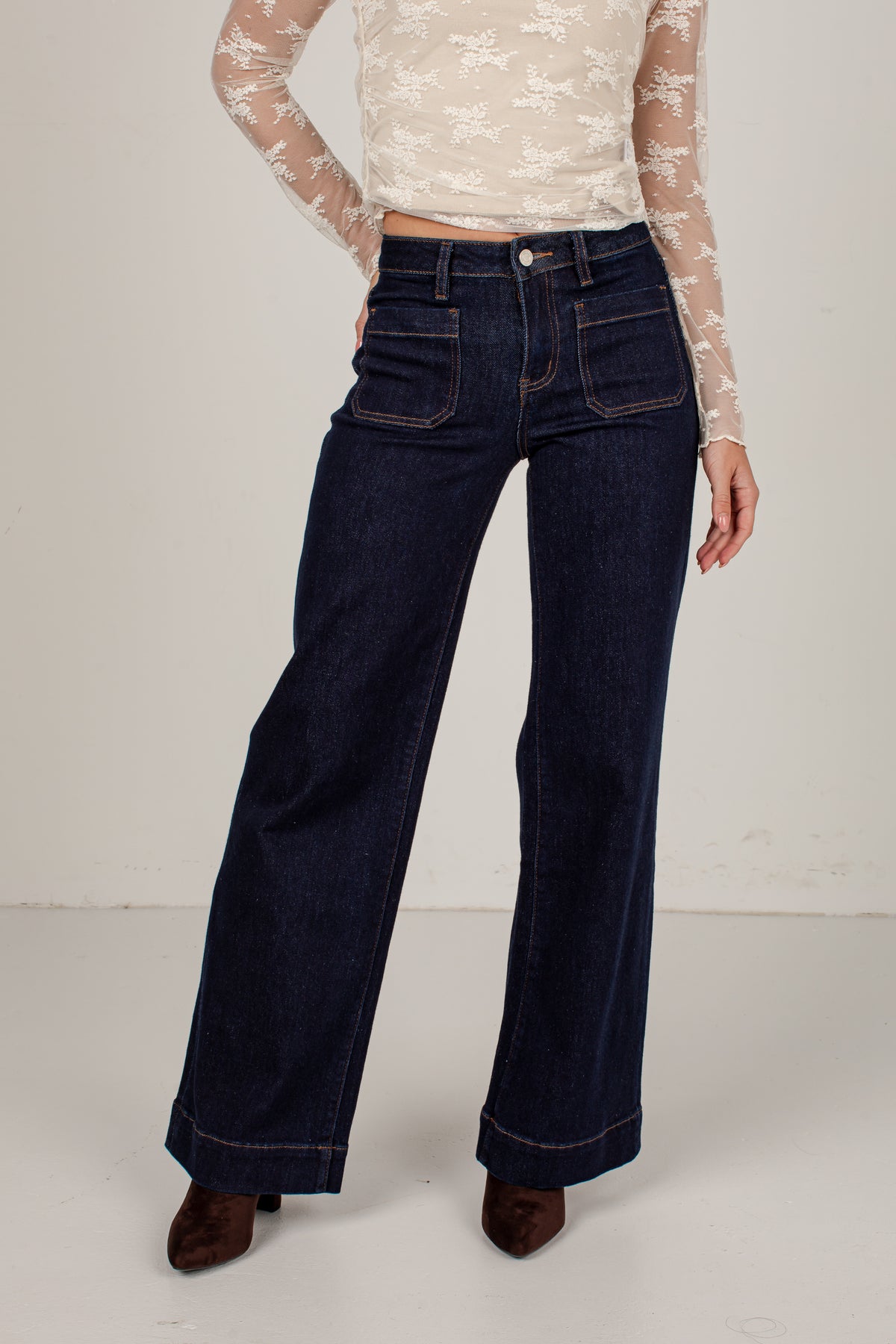 A model wearing dark blue wide-leg denim jeans, a cream lace long-sleeve top, and brown pointed-toe heels, standing against a plain off-white studio background.