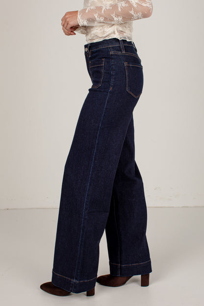 A person shown from the waist down wearing dark blue wide-leg jeans, a white lace long-sleeve top, and brown heeled shoes against a plain off-white studio background.