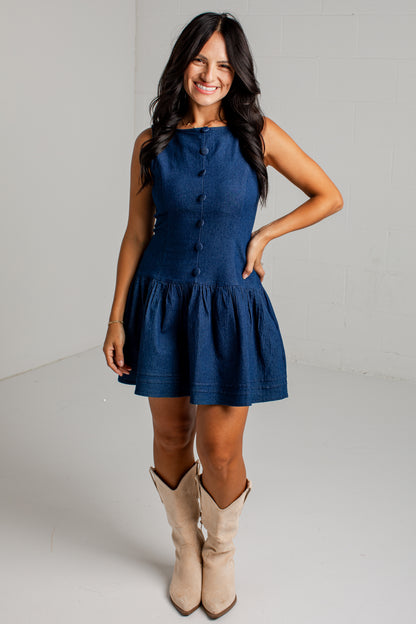 A woman modeling a sleeveless dark blue denim mini dress with a buttoned bodice and flared skirt, paired with beige cowboy boots, against a white studio background.