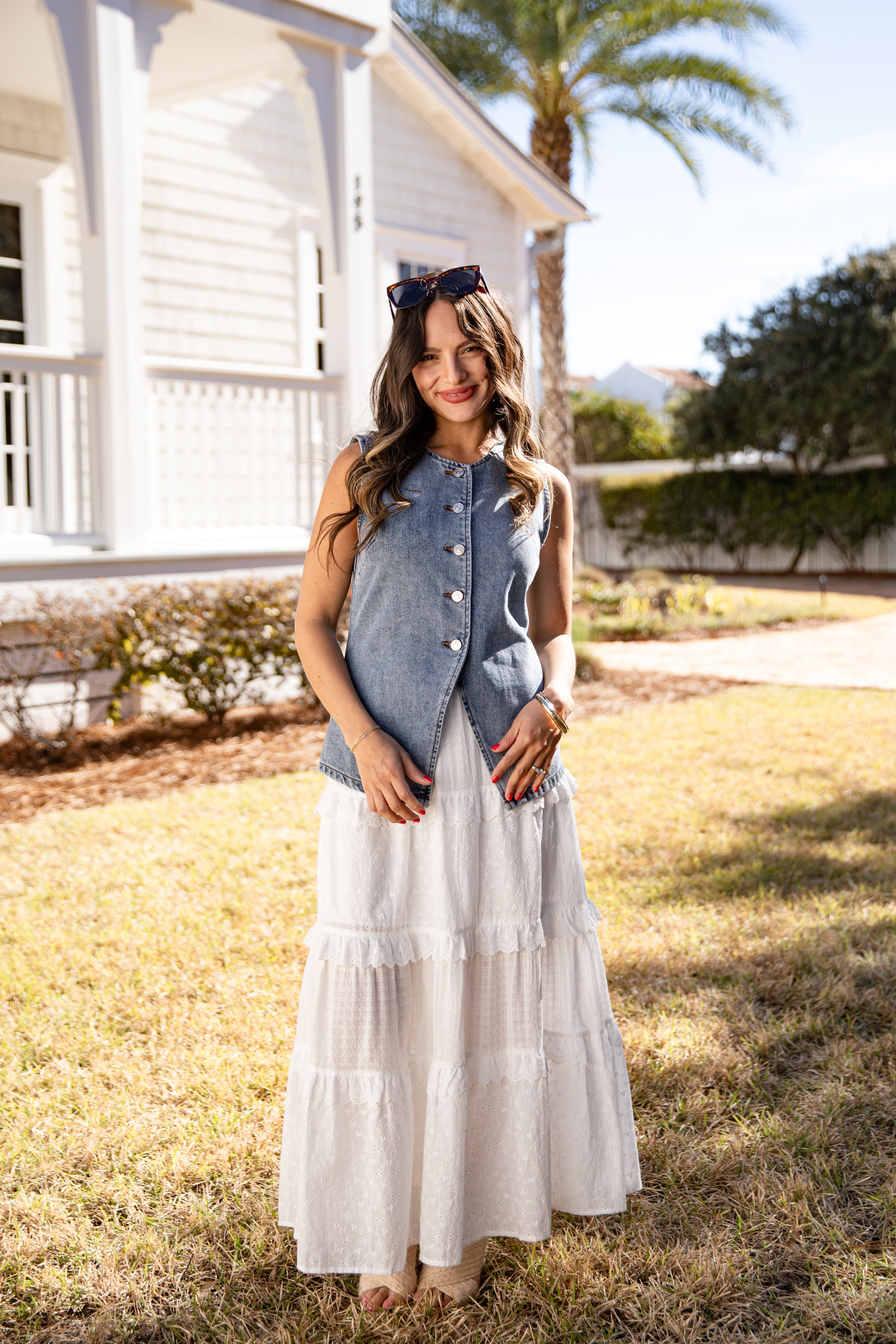 Woman in a denim vest and white dress standing in a grassy area with a house and palm tree in the background.