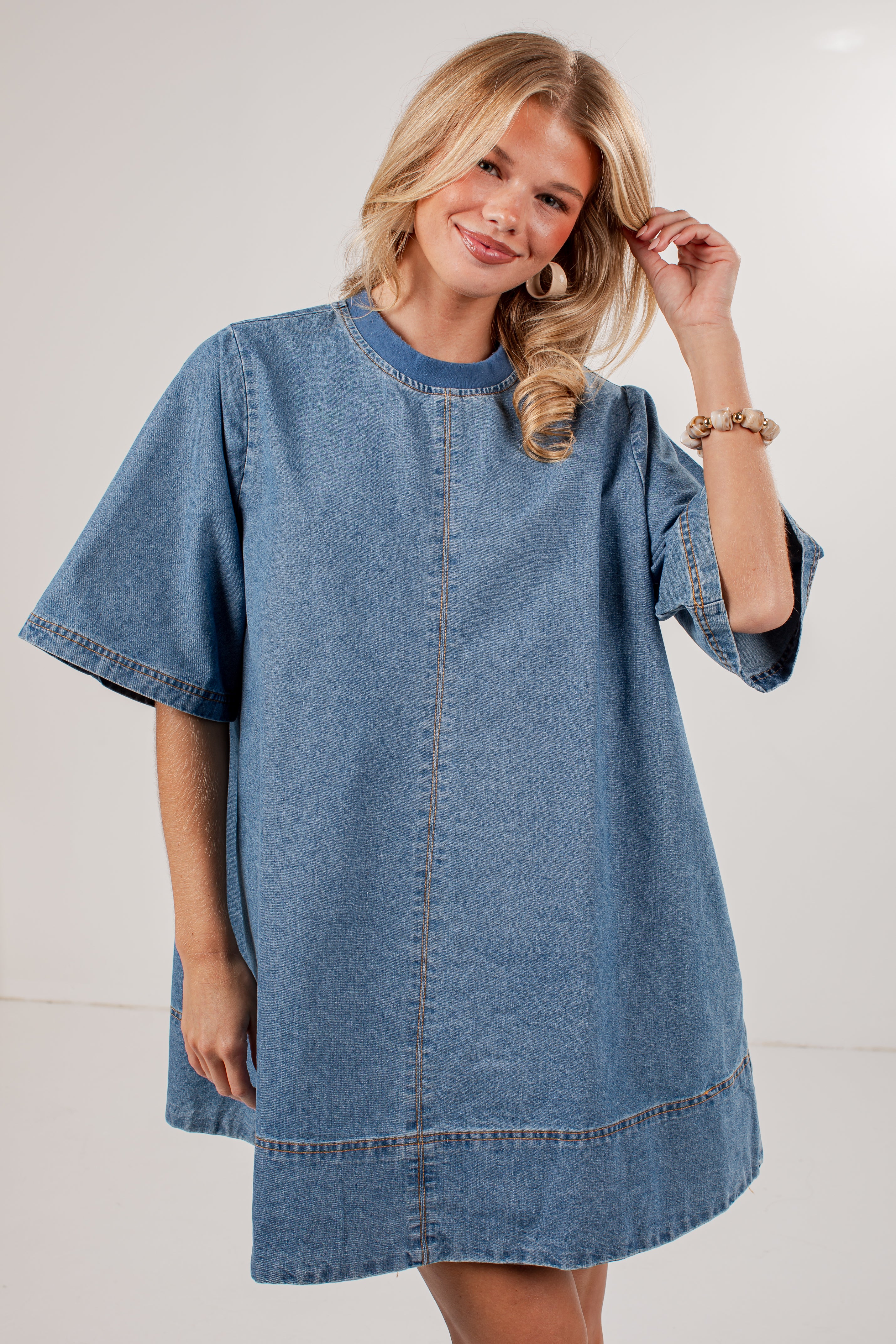 A smiling blonde woman wearing an oversized blue denim tunic dress with short wide sleeves, a round neckline, and contrast stitching, photographed against a plain light gray studio background.