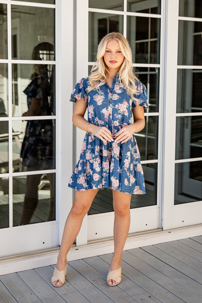 Woman in a blue floral dress standing in front of glass doors.