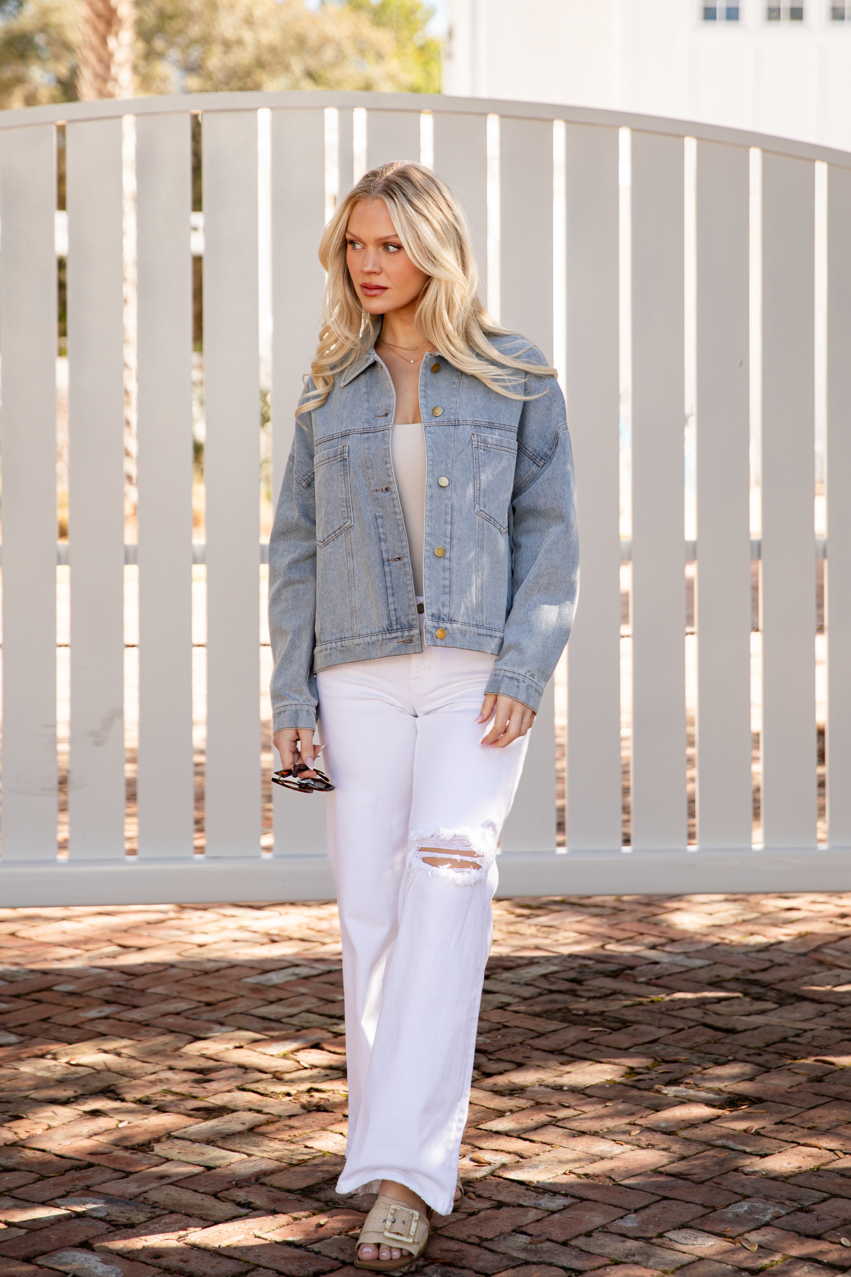 Woman wearing a denim jacket and white pants standing in front of a white fence.