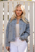Woman wearing a denim jacket and white pants standing in front of a white fence.