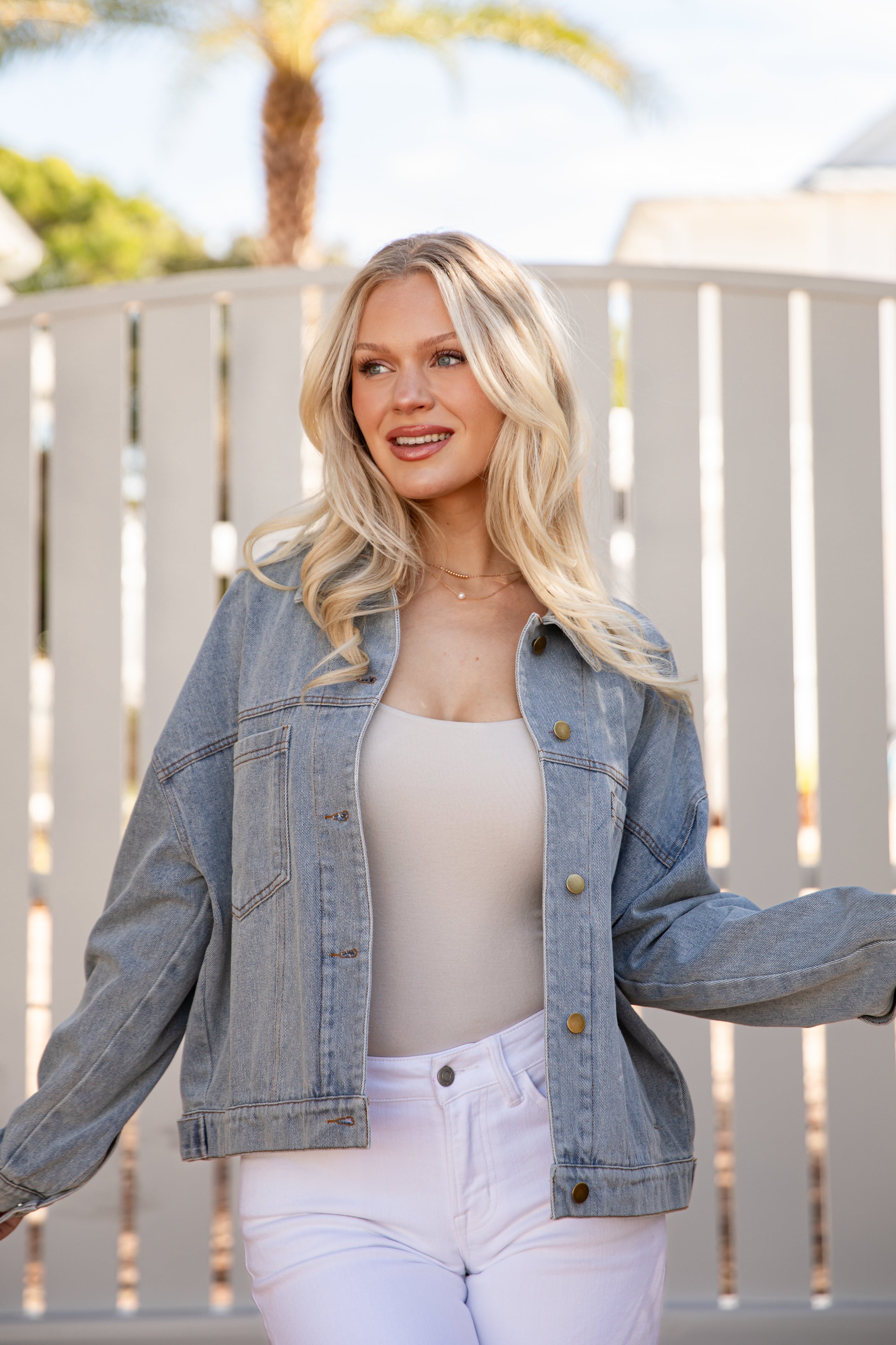 Woman wearing a denim jacket and white pants standing in front of a white fence with palm trees in the background.