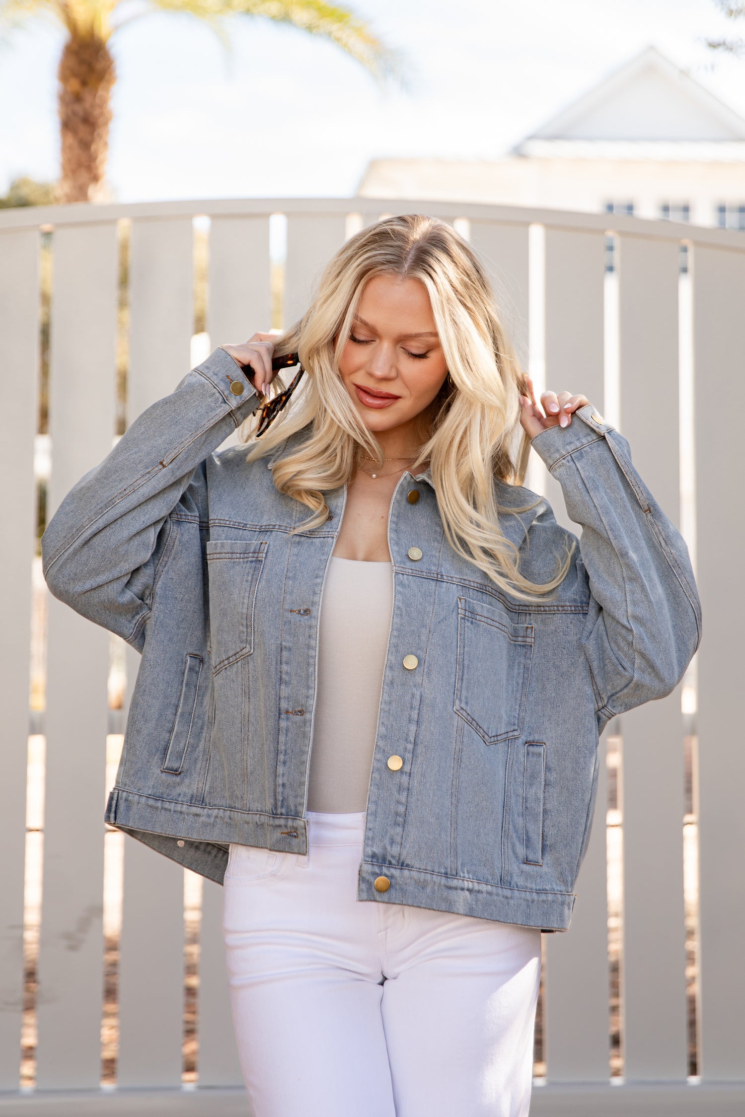 Woman wearing a denim jacket and white pants standing in front of a white fence.
